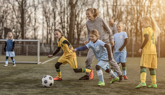 Fußball-Trainerin und mehrere Mädchen rennen lachend einem Fußball hinterher. Sie tragen Sportkleidung. Im Hintergrund steht ein weiteres Mädchen im Tor. 