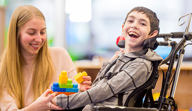 Eine junge Frau kniet neben einem Jungen im Rollstuhl, der in die Kamera blickt. Sie hält seine Hand. In der anderen Hand hält sie ein Stapel-Spielzeug. Beide lachen.